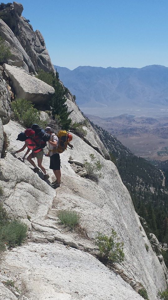 Three climbers wearing backpacks carefully cross a narrow granite ledge high above the valley.